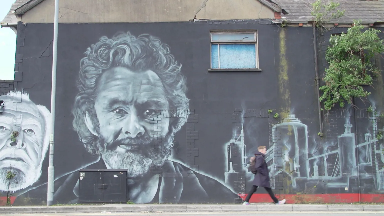 The photo is of a large two-storey mural of Michael Sheen in black and white, on the wall of a slightly rundown property. Next to the portrait of him is an image of the steelworks. There is one upstairs window with blue curtains and some greenery growing in a crack in the masonry. In front of this Jon can be seen walking along the pavement in a grey coat witha. rucksack. 