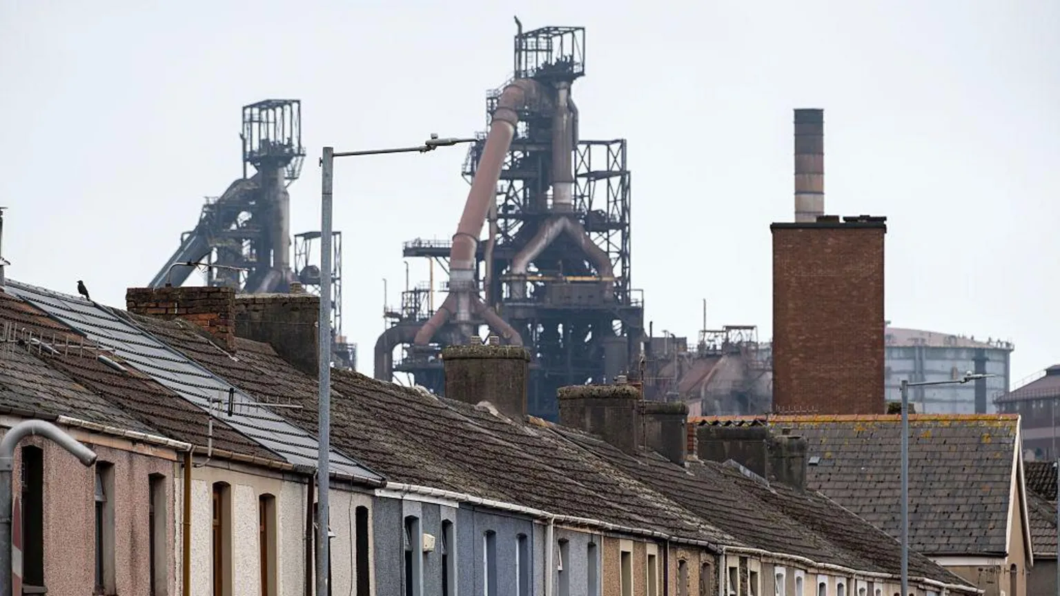  The first floor and roofs of a row of a street of colourful terraced homes can be seen across the lower half of the picture. The skyline behind them is dominated by a grid of pipes and metalwork which make up the two blast furnaces at Tata steel. 
