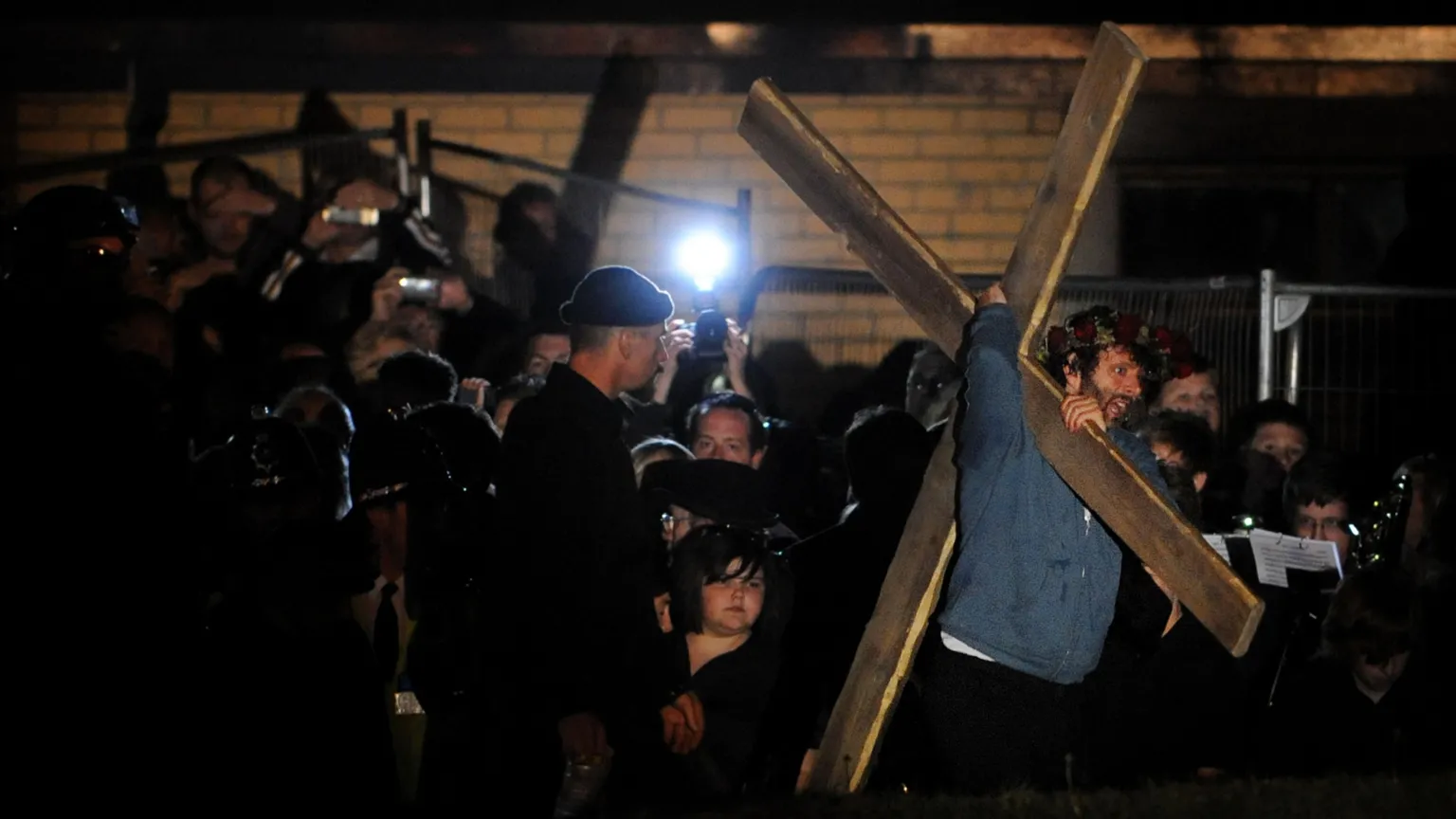  It's night. In the right of the photo Michael Sheen is dragging a cross, which is sat on his shoulder. He is clearly uncomfortable. Looking on is someone in black clothes and a black beanie. He gives off a menacing air. In the background is a huge crowd of spectators and media with cameras and lights to be seen. Behind them is the wall of a red-brick community centre. 