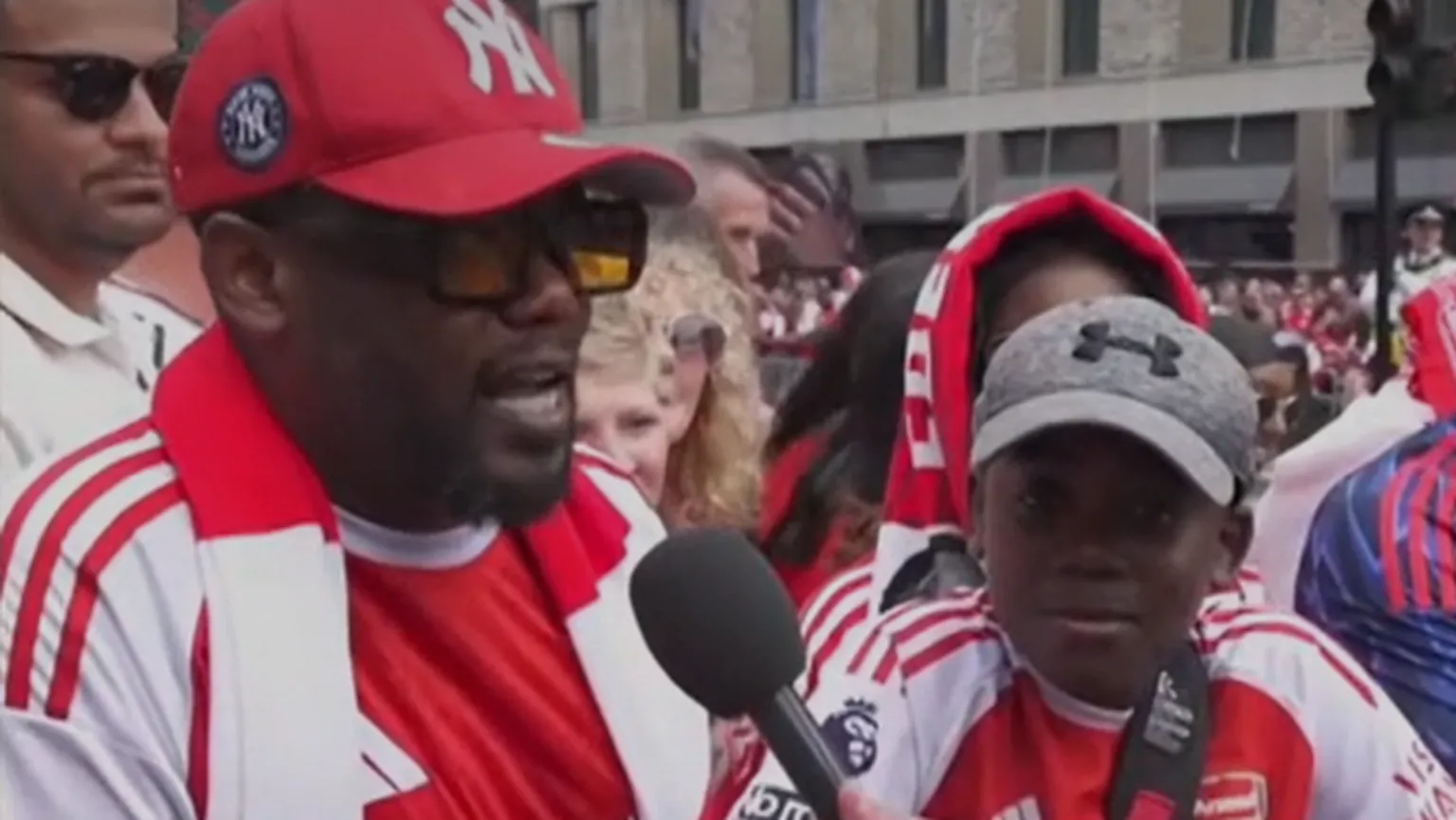 Joe speaking into a microphone, wearing an Arsenal T-Shirt and scarf, flanked by his son who is smiling past the camera