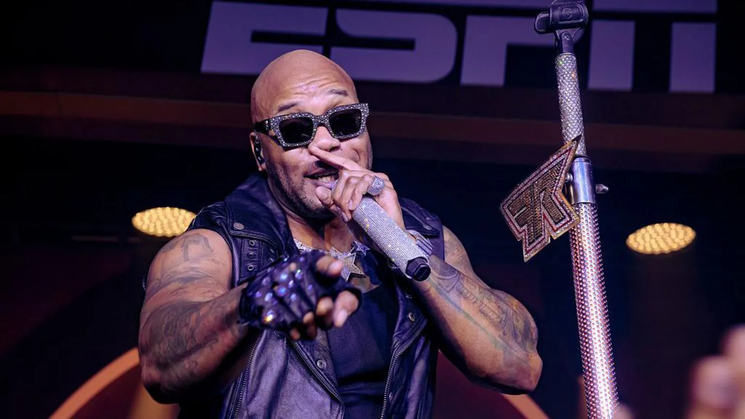 A man wearing rhinestone-studded sunglasses sings into a rhinestone studded microphone while he points with his right hand