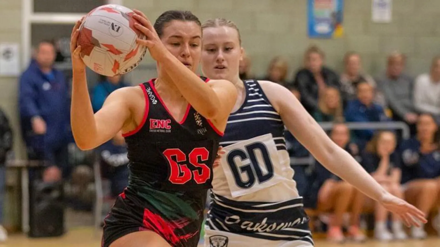 CRY Kaitlin, dressed in black and red sports kit, is playing in a netball match and is holding the ball. An oponent, dressed in white and black, is chasing her.