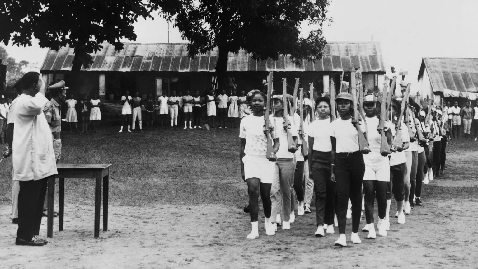 AFP via Young female militia of the civil defence - most dressed in white and with white socks and trainers - hold what look like rifles against their shoulders as they parade during military training in Enugu in August 1967. To the side a man in a white coat and dark trousers salutes as two other soldiers look on. It is a black and white archive photo.
