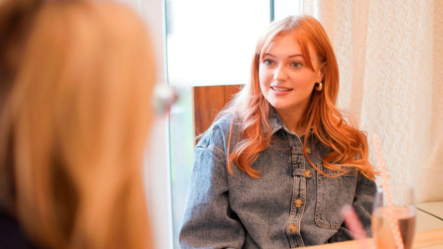 Chloe, a woman with long, red hair and who is wearing a button-up denim jacket, sits at a table talking to the BBC's Emma Barnett, who is visible from behind close to the camera, while light streams in behind her from what looks like a patio door.