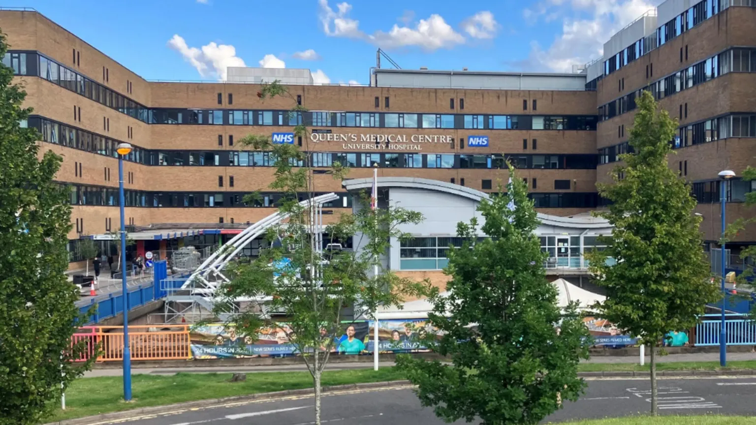 Front entrance of Queen's Medical Centre in Nottingham. A four-storey high building. Another, smaller, more modern building is in front - with a curved roof. There are small trees in the foreground, and the sky is blue with white fluffy clouds.