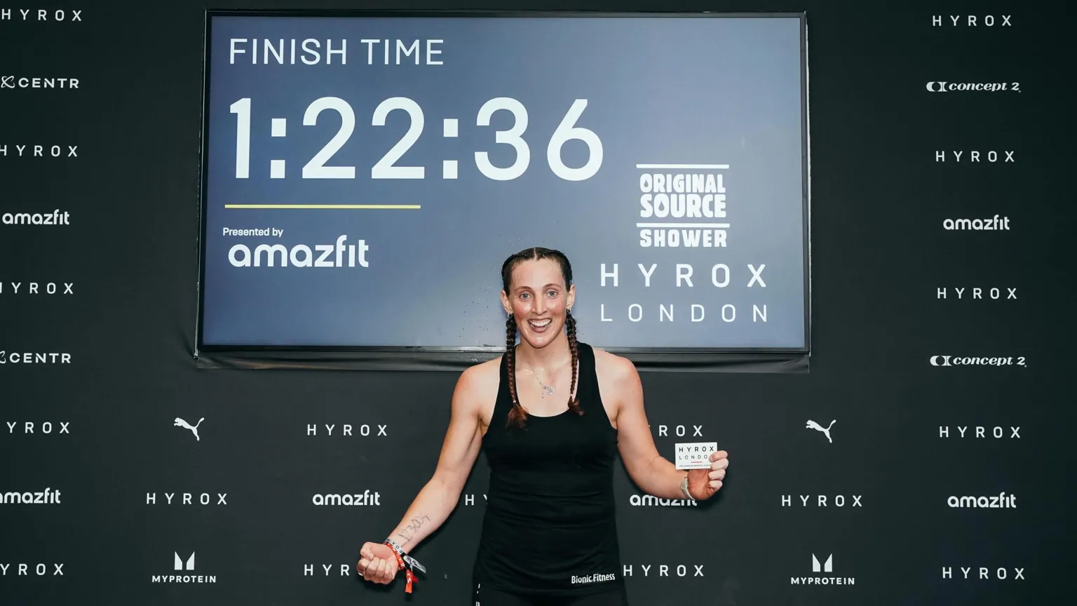 Beth Littlewood A woman wearing all black sports kit and her brown hair in two plaits. She is holding a small card in one hand with the words Hyrox London on it. The other hand is clenched in a fist. She is smiling and looking at the camera. Behind her is a board with the Hyrox London logo in one corner and then the words: