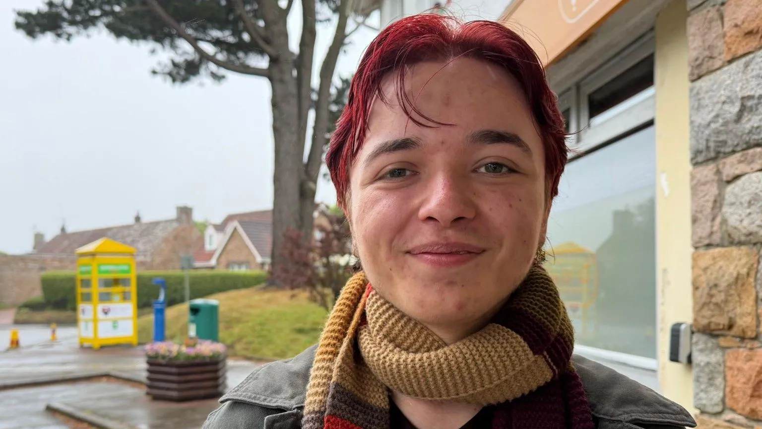 Toby Cahill Le Brocq has red hair and a beige scarf. He is wearing a black jacket. 
