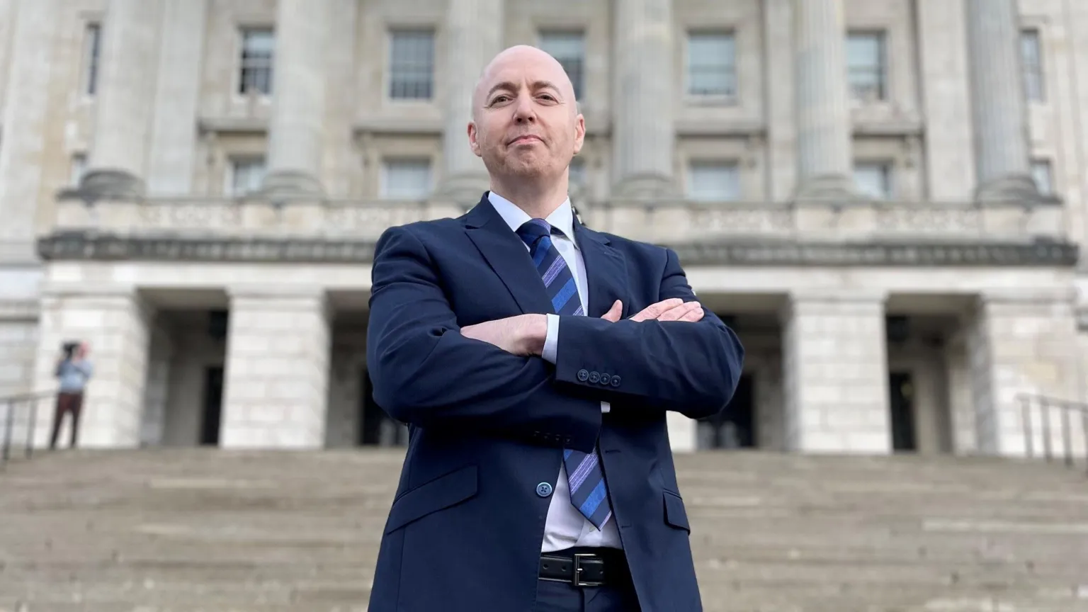 David Young/PA Wire Jon Burrows. a man with a shaved head, stands on the steps of Stormont with his arms folded, looking down at the camera. He is wearing a navy suit, a white shirt and a navy and mid-blue striped tie.