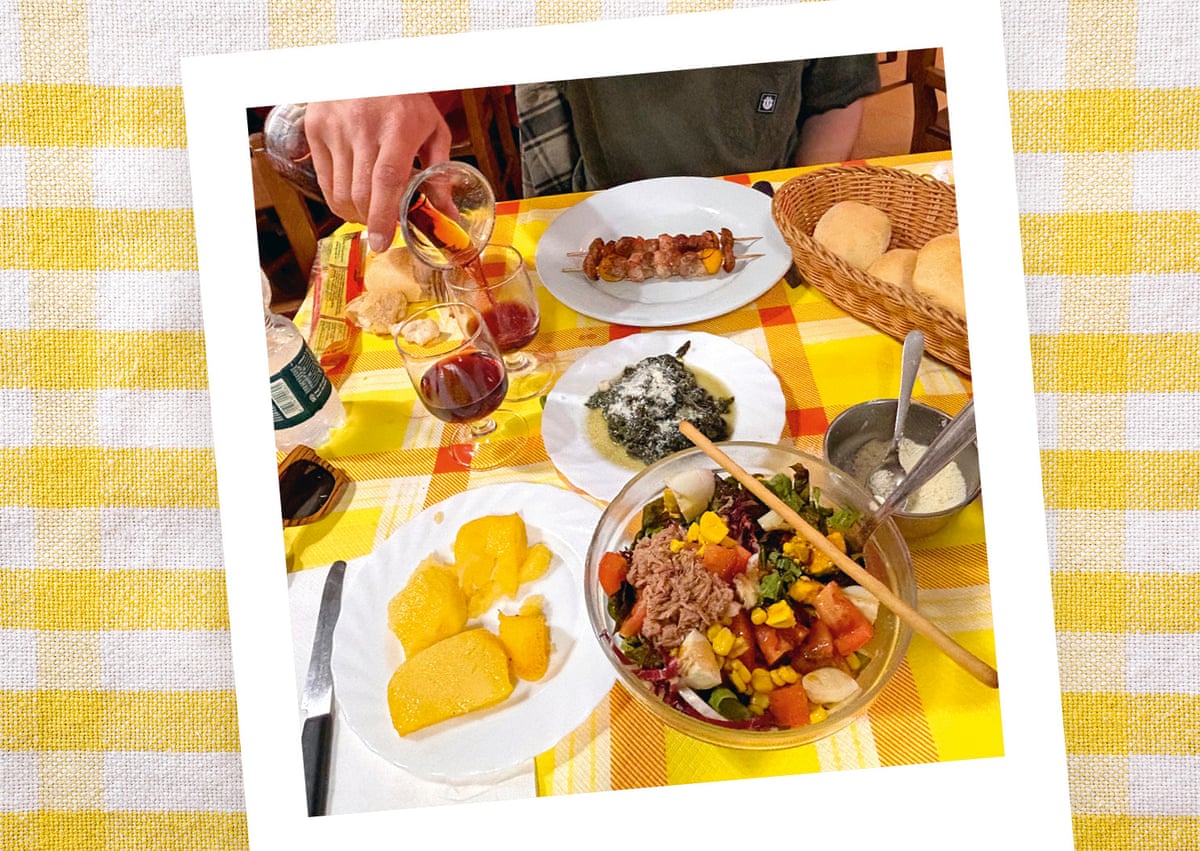 A table spread with salad, grilled meat skewers, polenta, greens, bread, and wine on a checkered tablecloth