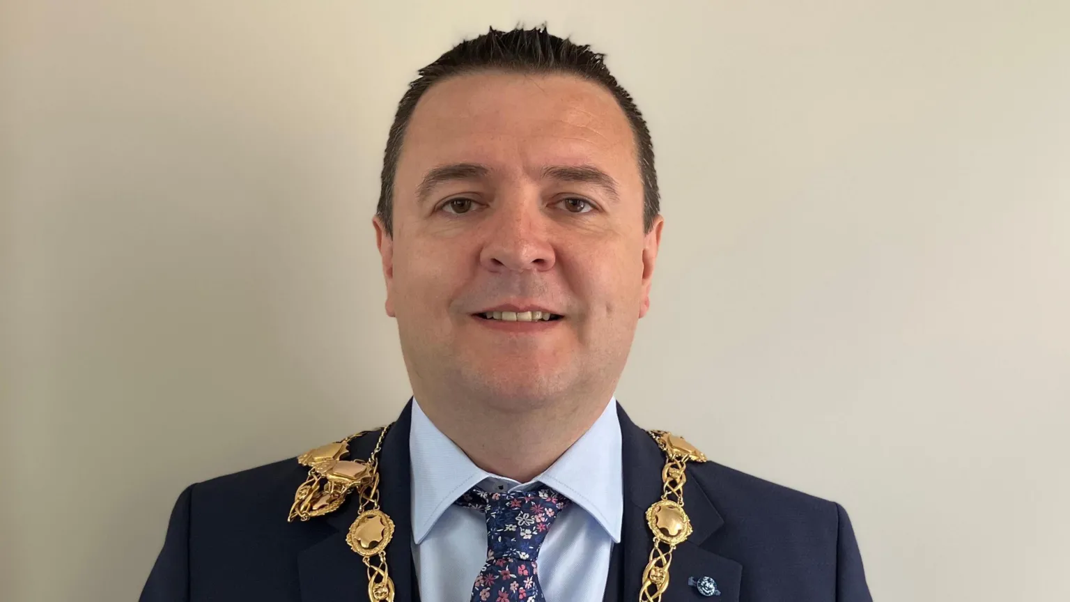Derry mayor Ruairí McHugh looking directly at the camera and smiling. He has very short, dark hair and is wearing a navy suit, pale blue shirt, navy patterned tie and his gold chain of office.