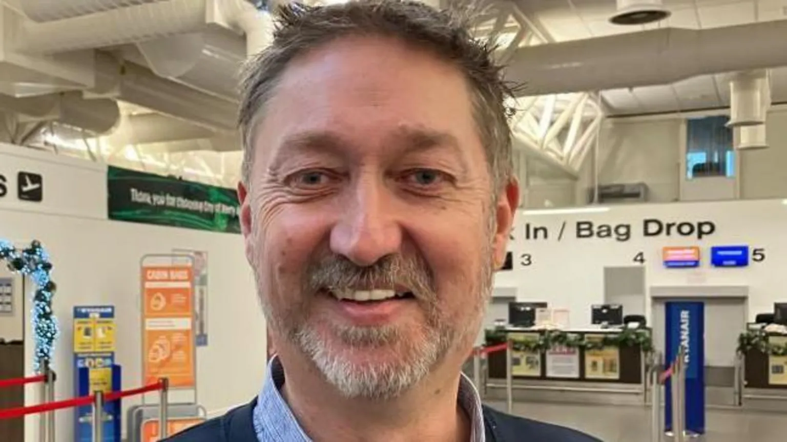 Steve Frazer has short brown hair and a beard. He is standing in the City of Derry Airport main lounge. 