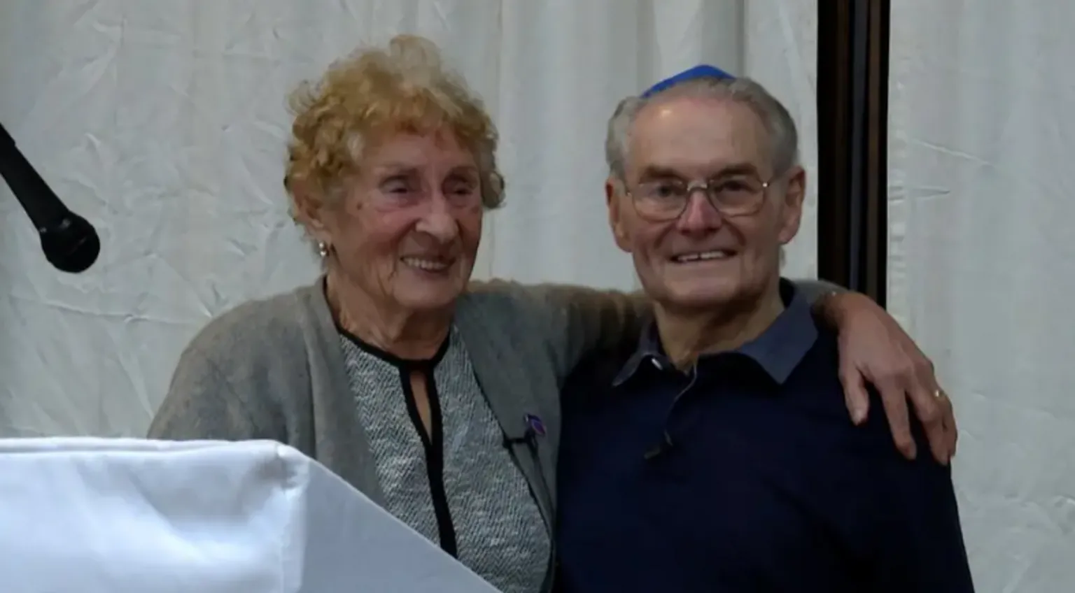 Susan Pollock and Tomi Reichental are standing side-by-side. Susan has curly red hair and is wearing a grey cardigan and top. Tomi Reichental is wearing a blue yamaka, a brimless, usually cloth skullcap traditionally worn by Jewish men, and a black sweater and navy shirt.