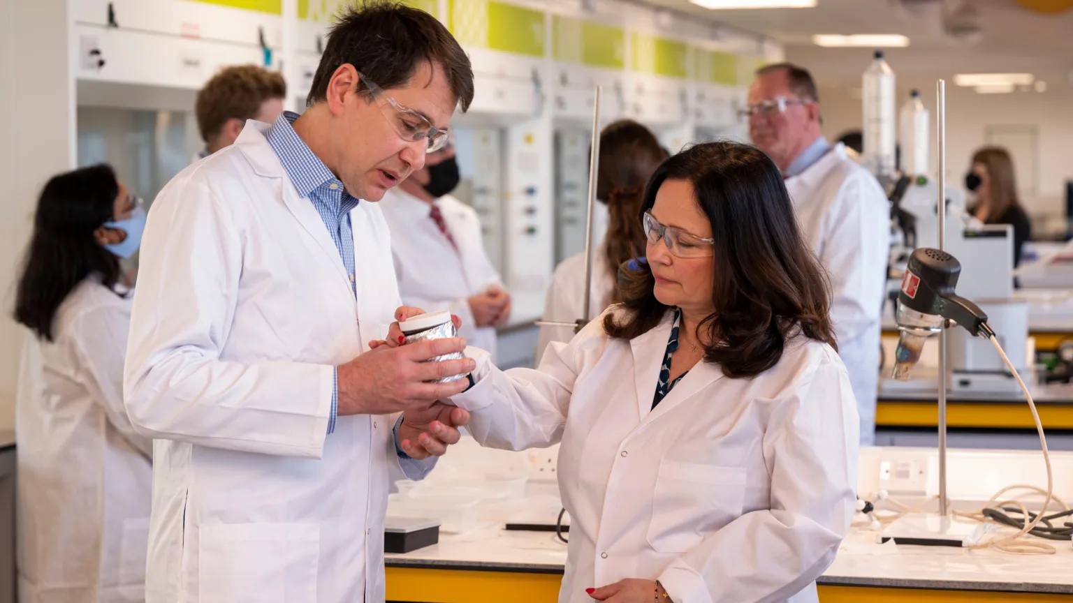  A dark-haired man and woman in white lab coats and goggles in a research lab