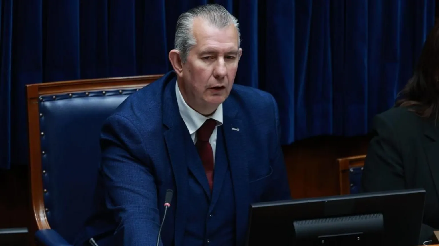  A man with grey hair is sitting on a wooden chair with navy leather. There is a navy curtain in the background. The man is wearing a navy three piece suit, white shirt and dark red tie.