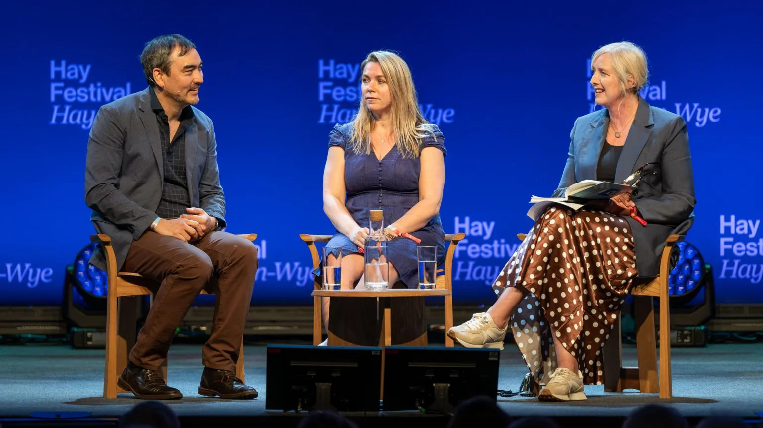 Hay Festival Sarah sits between Tim Wu on the left and Carole Cadwalladr on the right on stage at hay. they are looking at each other with tim and carole smiling and Sarah looking expressionless. 