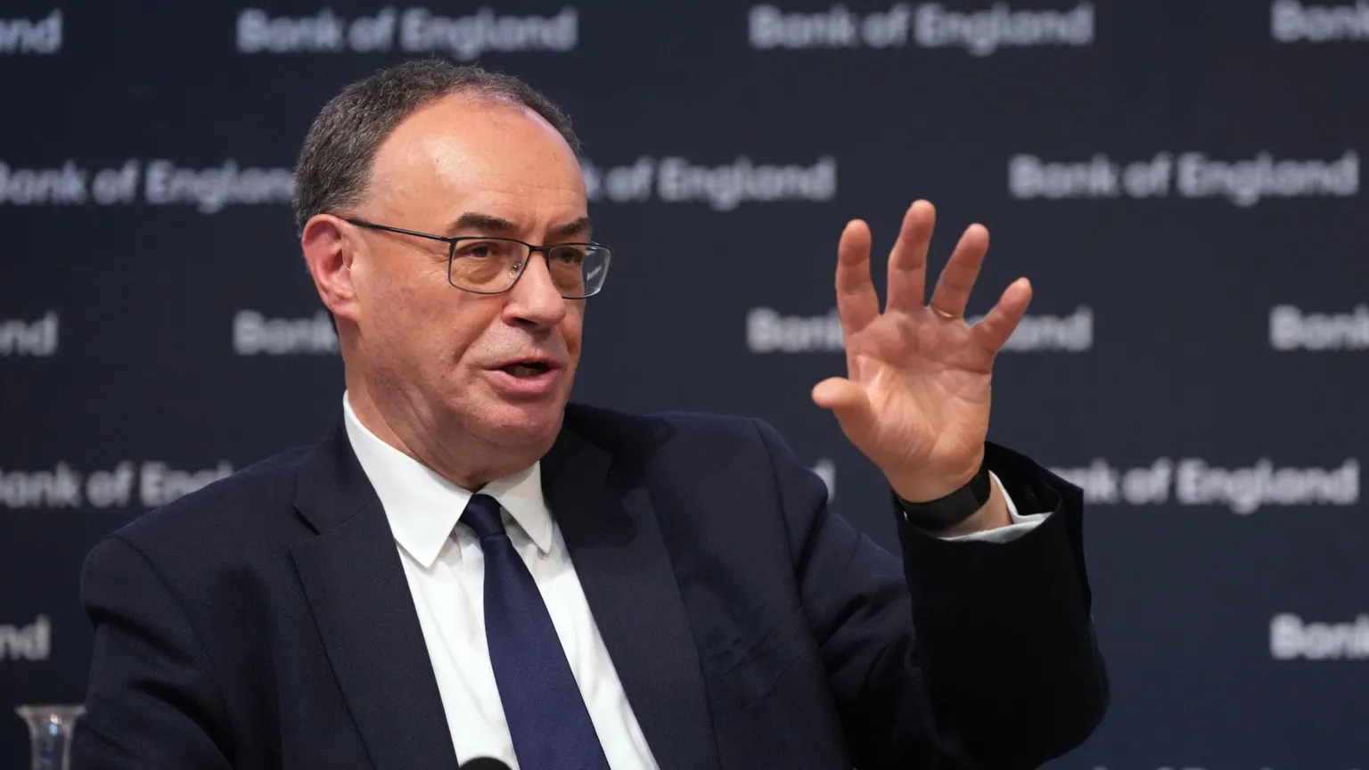 Governor of the Bank of England Andrew Bailey sits on a stage, addressing the audience and gesturing with an open hand. He wears a dark suit, white shirt, dark tie and glasses.
