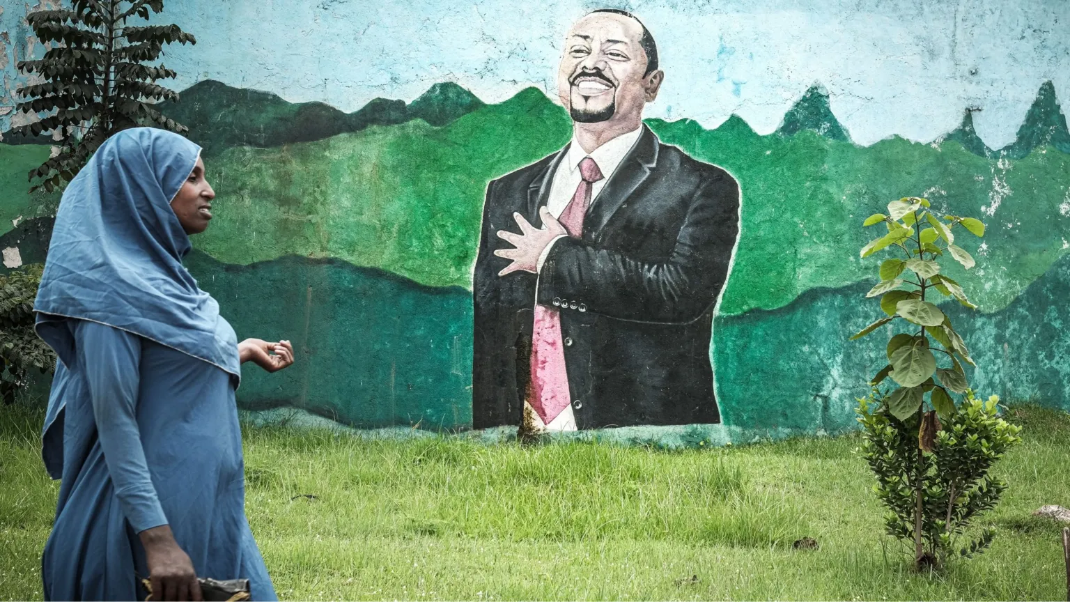 AFP via A woman in a blue dress and headscarf walks past grass in front of a wall with a mural of Abiy Ahmed. He is depicted in a black suit, white shirt and red tie holding his arm up over his heart.