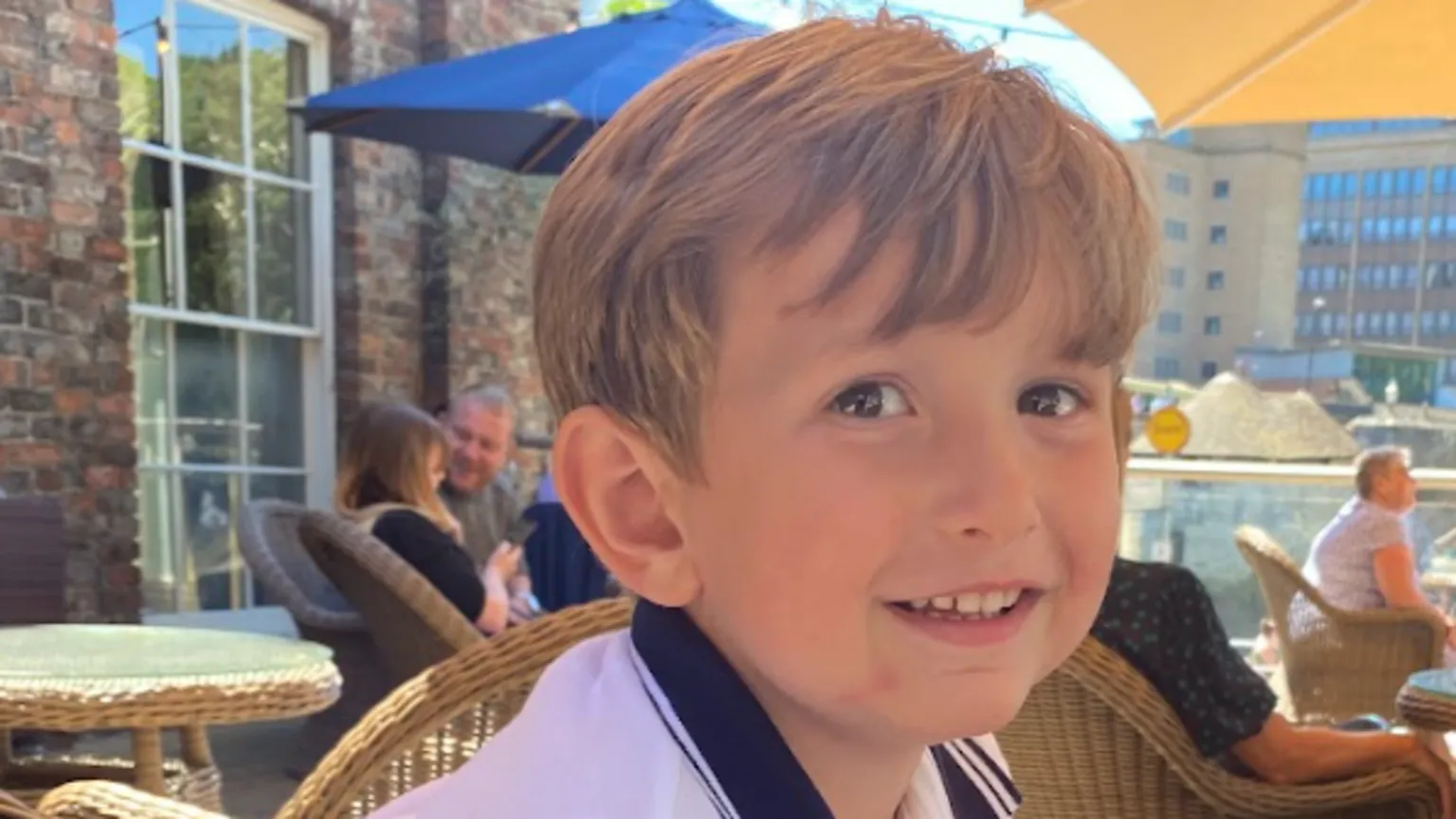 Family Handout A boy with blonde hair smiles at the camera He is wearing a white polo shirt with a black collar. The photo is taken outside in a restaurant with people chatting at different tables.