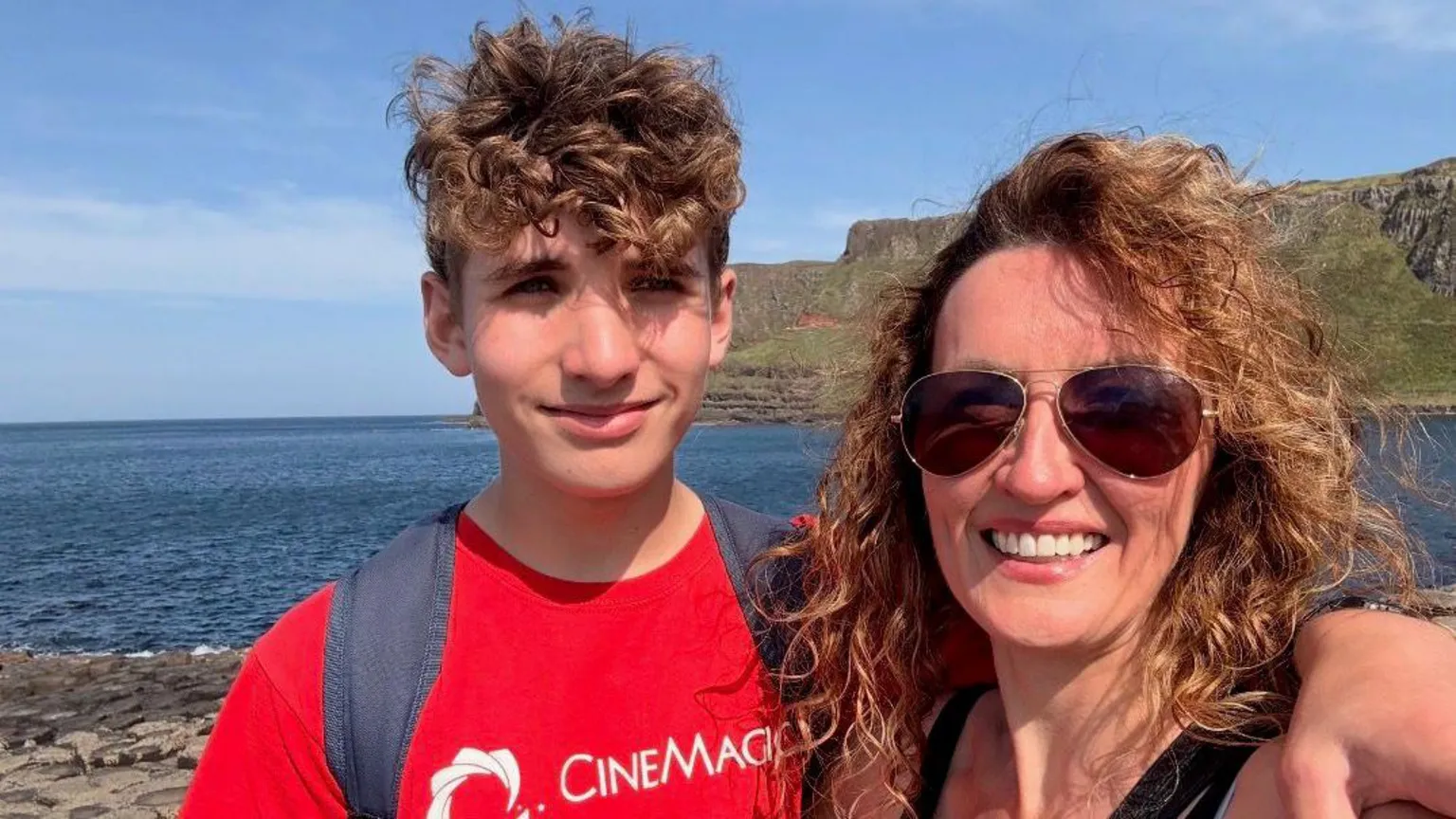 Family Handout A photo of a boy (left) and a woman (right) The boy is wearing a red tee shirt with the words 'cinemagic' written in white. He has brown curly hair and is smiling at the camera. He is also wearing a black backpack that you can see the straps over his shoulders. The woman is wearing brown sunglasses, with brown curly hair and is smiling at the camera. In the background there is the cliffs and sea.