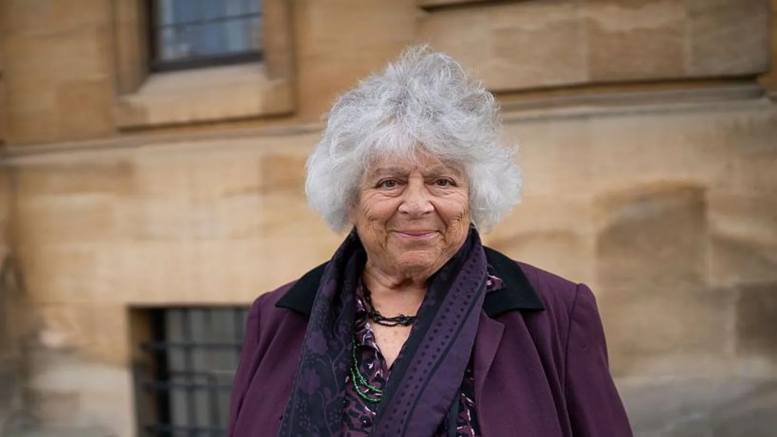  Margolyes smiles as she stands in front of a brown building at the Oxford Literary Festival 2025. She wears a purple blazer, a purple blouse, a purple scarf and has short grey curly hair.