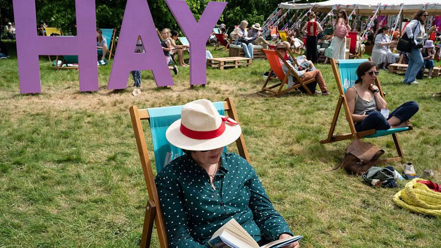  A person in a white brimmed hat and shirt is seen sat reading a book, in front of a large purple sign reading 