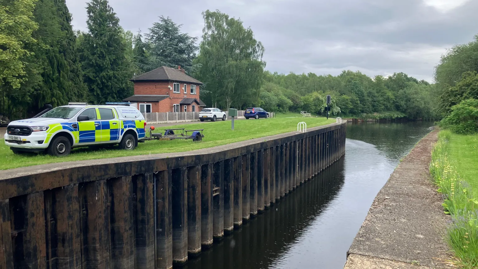 Phil Bodmer/BBC Police search vehicle with a trailer next to a river, on a verge of grass. There is a house in the background.