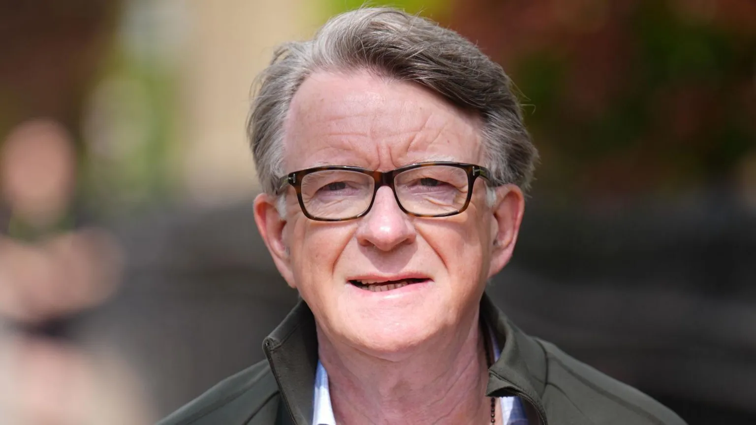 Peter Mandelson has short greying hair and wears dark glasses. He wears a shirt, grey jacket and appears to be outside