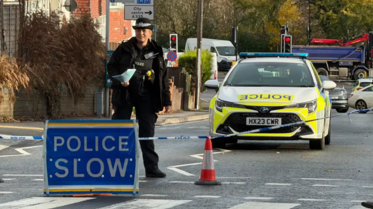 A police cordon with a Police Slow sign, blue tape, a police officer and a police car blocking the road. A pedestrian crossing can be seen in the foreground with other vehicles including lorries in the background.