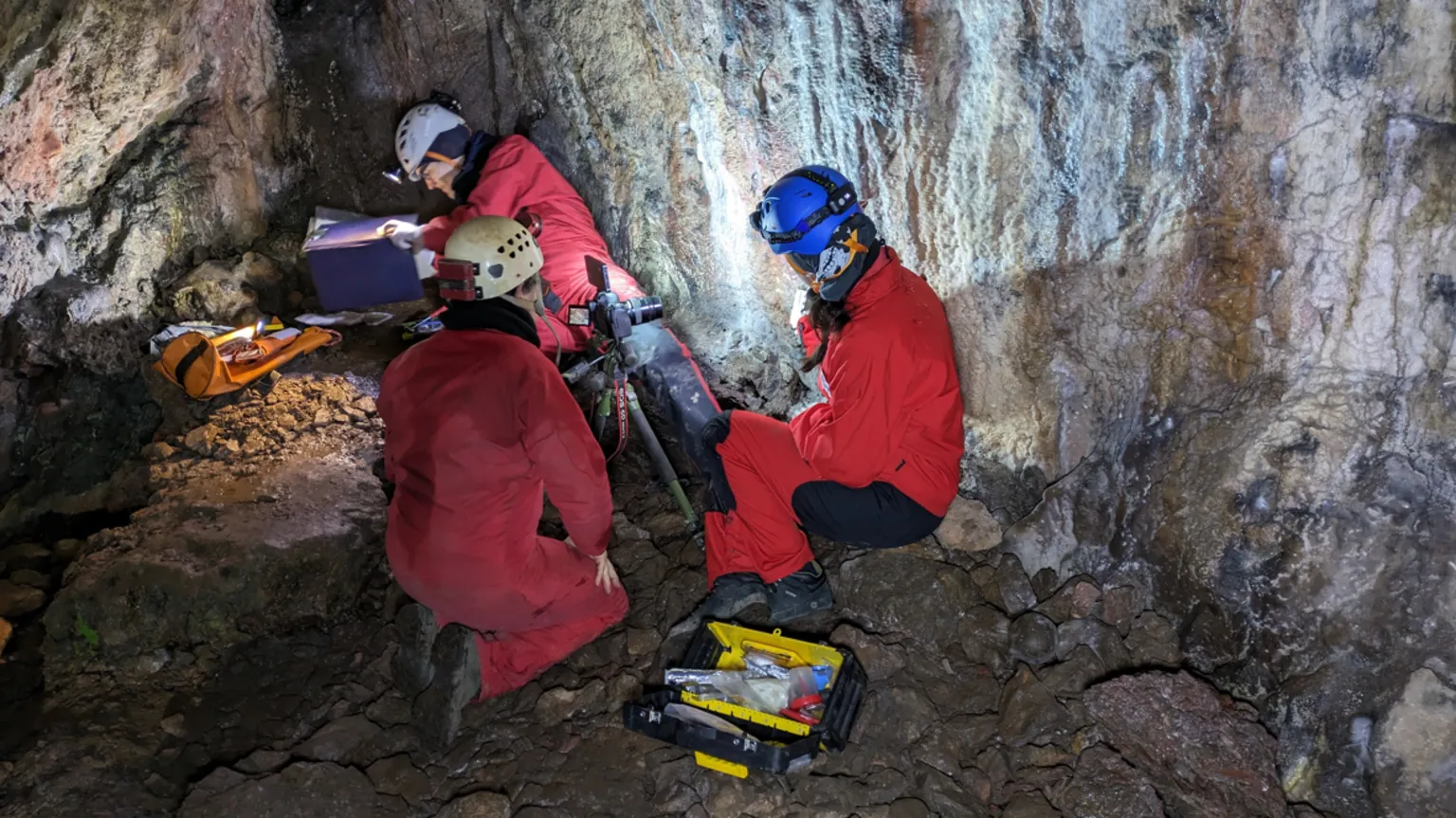 George Nash Three people in red boiler suits sit on a cave floor with the rock walls behind them. They wear helmets with torches on the front. A tool box lies on the ground by them.