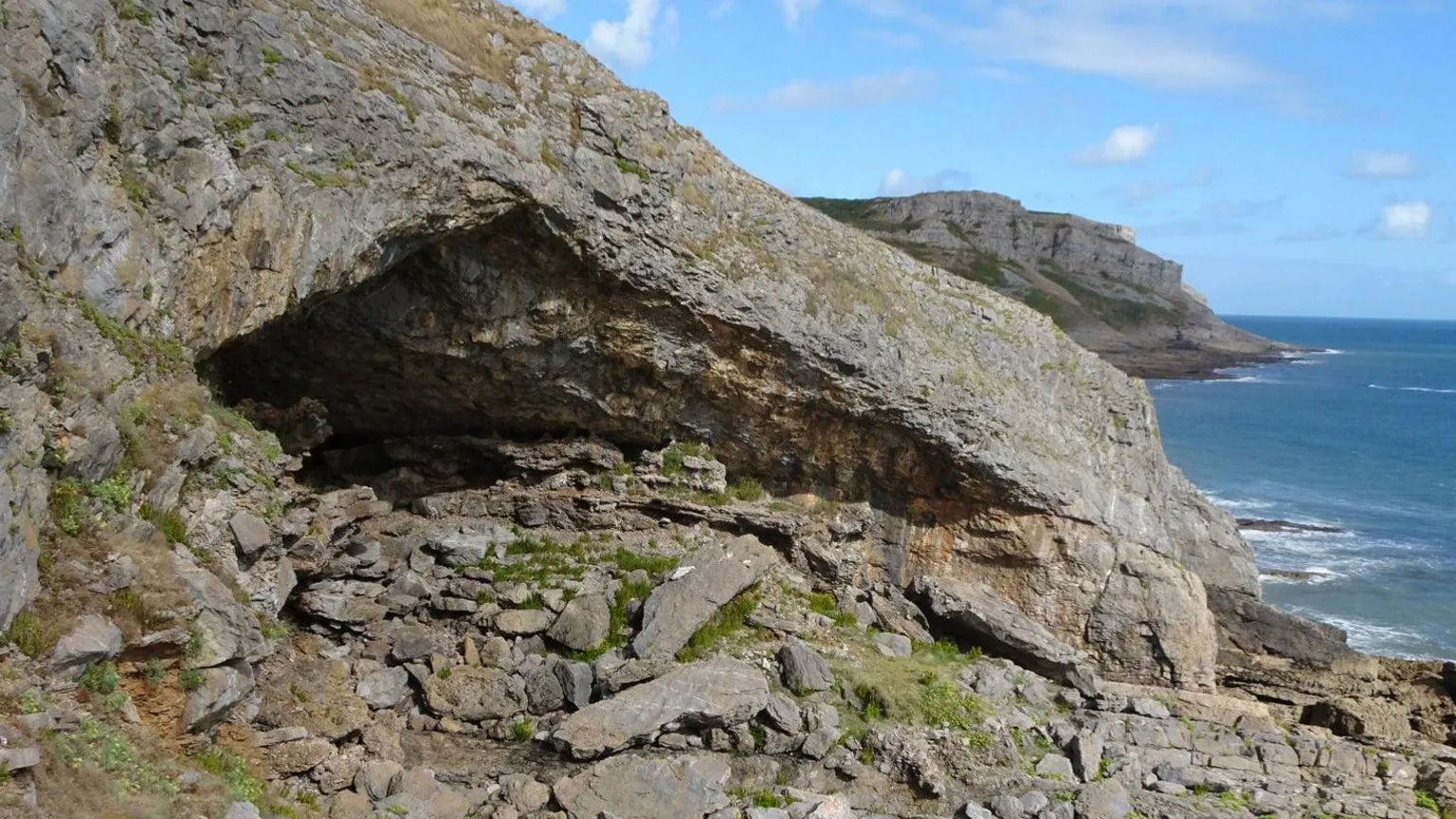George Nash A general view of Bacon Hole cave. Its opening is in the shape of a triange, with the sea and more cliffs behind it, with the blue sky beyond. There are small rocks covering the ground in front of the cave entrace.