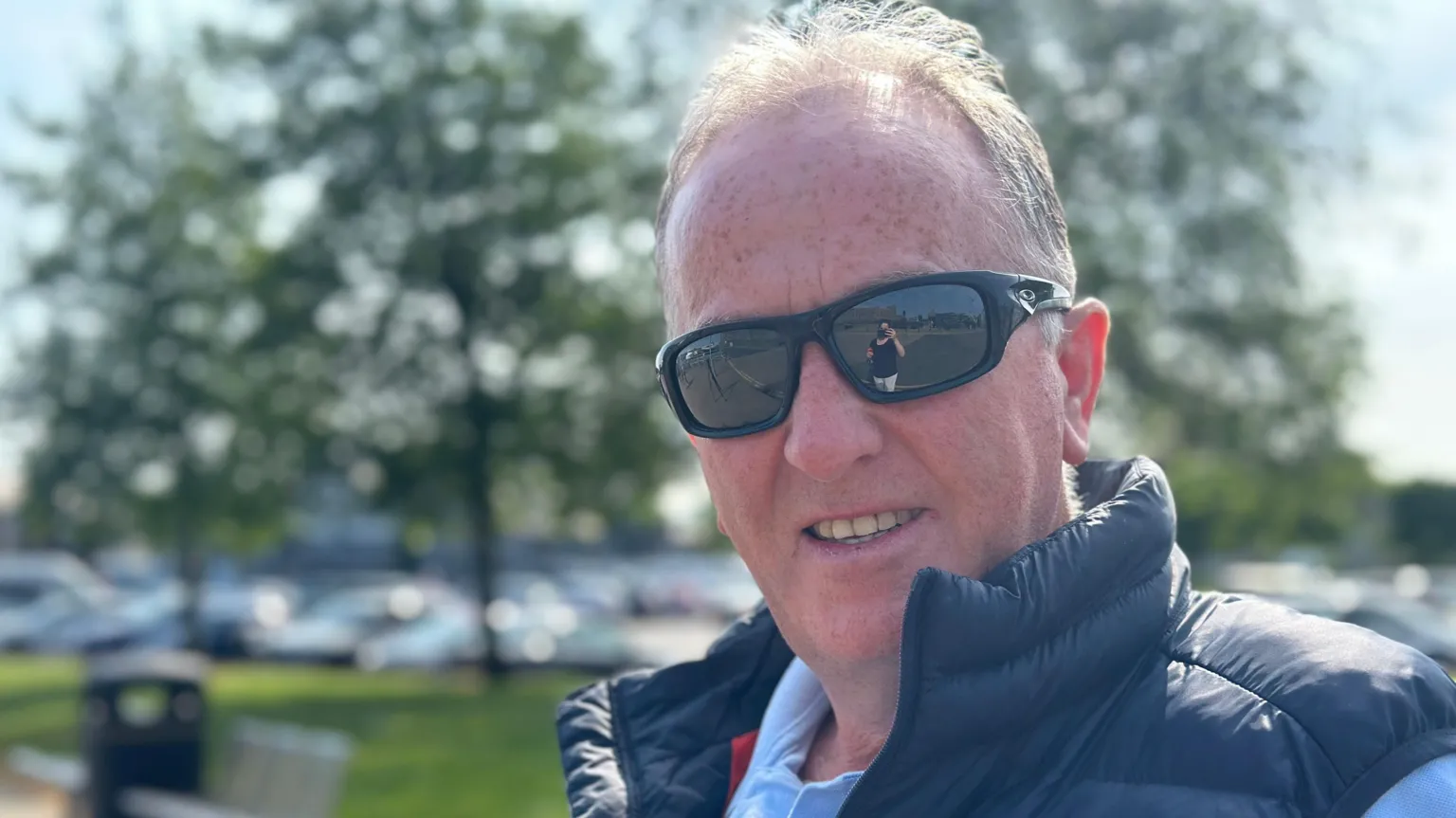 Michael looking at the camera. He is standing outside on a sunny day. He is wearing a navy jacket, blue shirt and sunglasses.