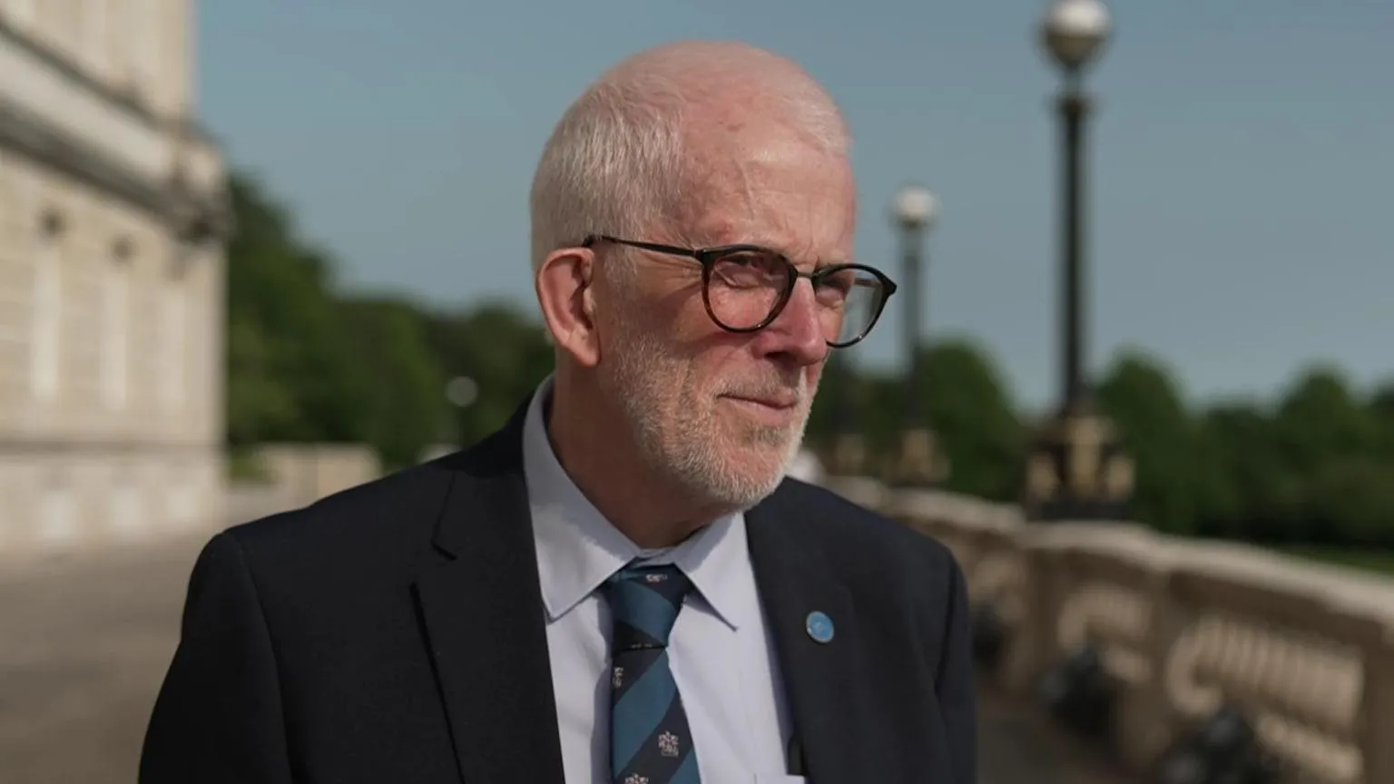 Stewart Dickson standing outside Stormont. He is wearing black-framed glasses, a black suit jacket, a striped tie and white shirt.