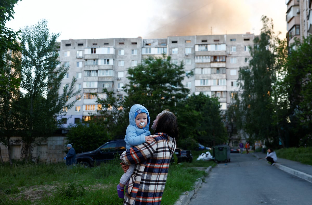 A woman carries a toddler as smoke rises above a Kyiv multi-storey building after Russia attacked, hitting apartment buildings