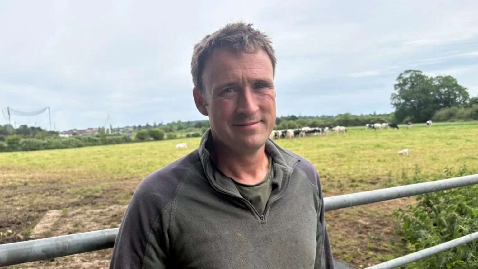 A man with short brown hair and a dark green fleece in standing in front of a gate, with a field of cows in the background.