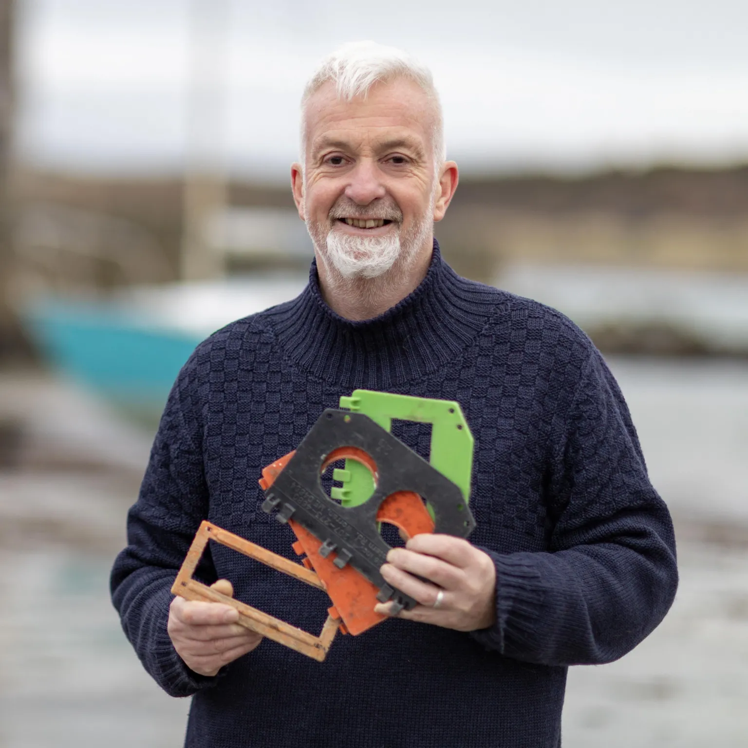 Mike Bolam Middle-aged man with grey hair and beard, holding a variety of plastic litter found on the shoreline. 
