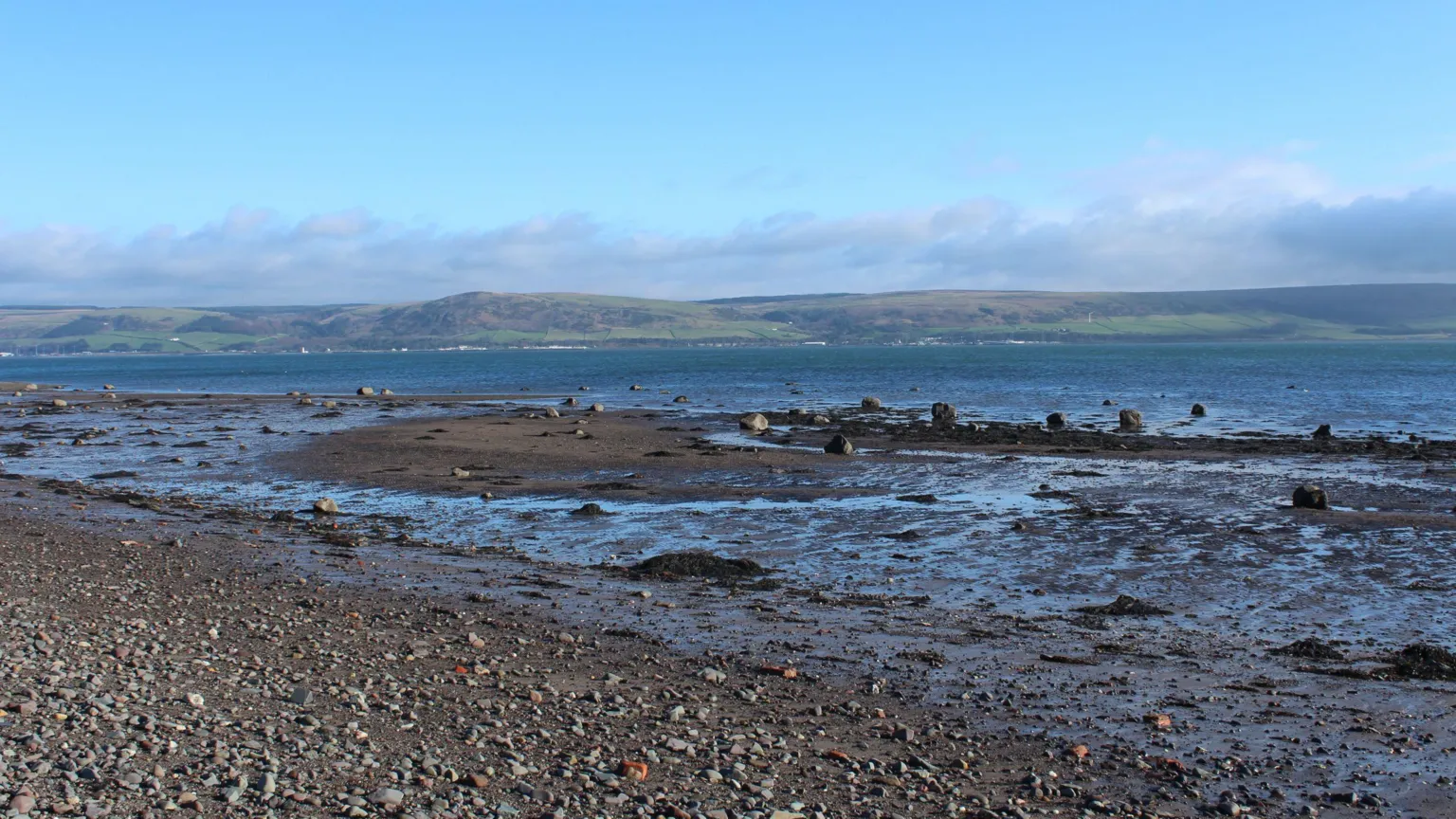 Billy McCrorie A Galloway shoreline with rocks and muddy sand looking out across the water to low, green hills in the distance