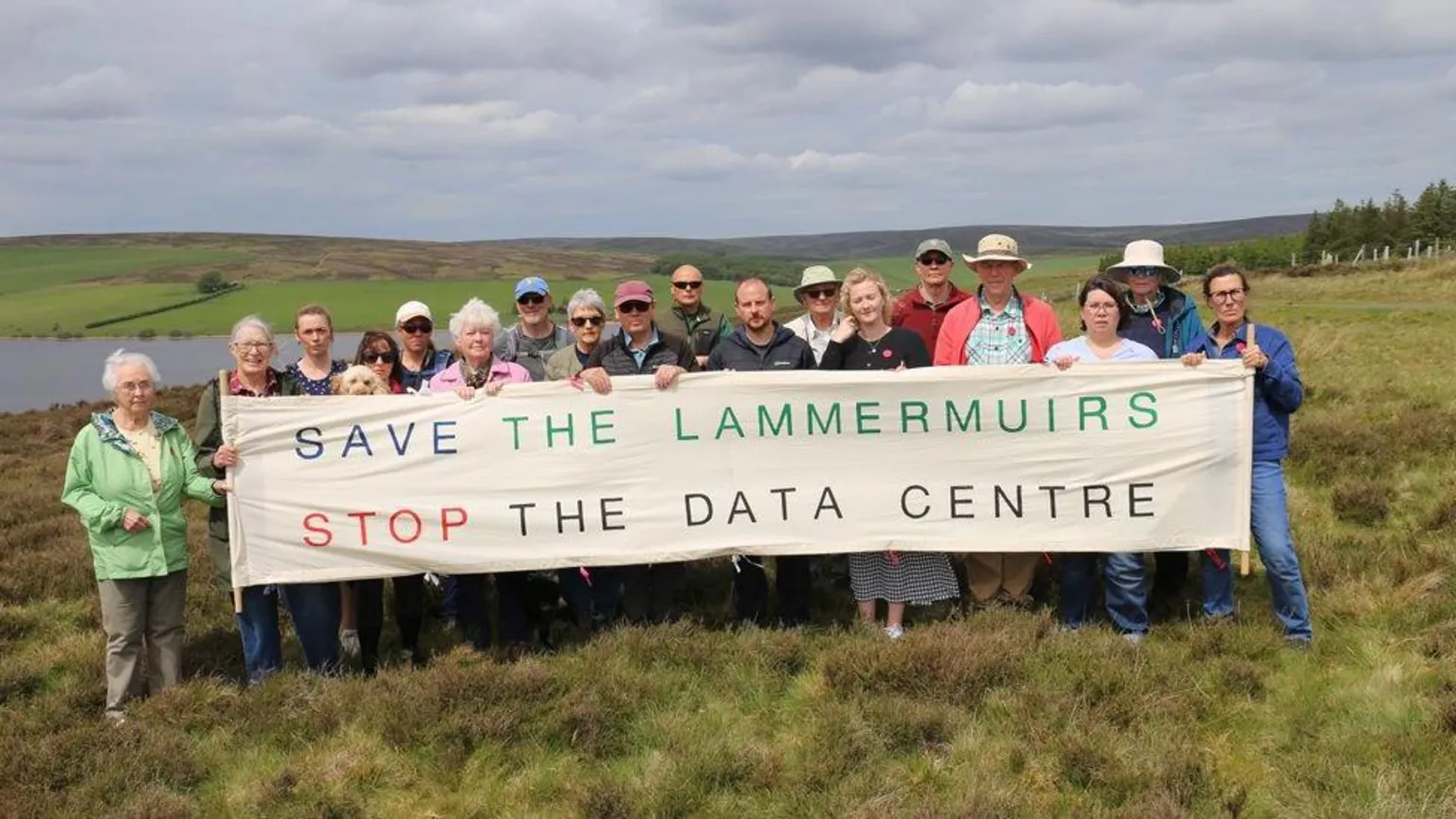 SLSDC A group of about 20 people standing behind a banner which says Save the Lammermuirs Stop the Data Centre. They are standing in rural countryside with a reservoir behind them.