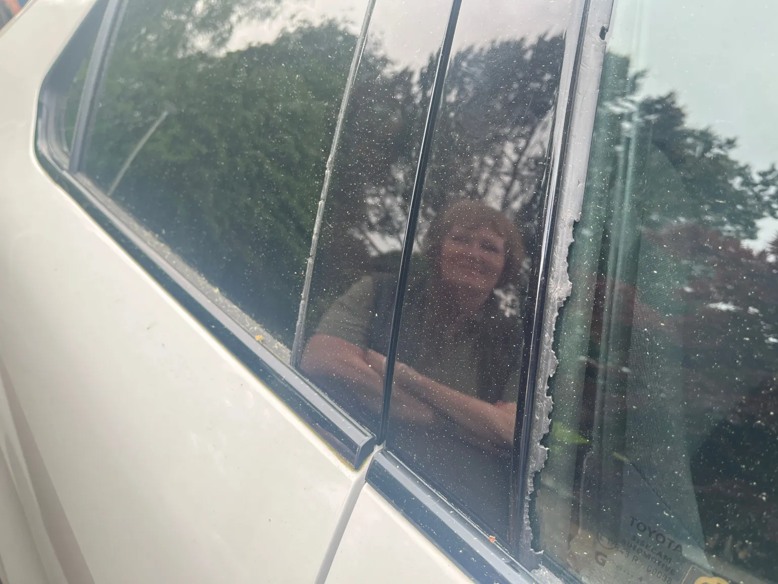 A photograph of the damaged car window seal. The reflection of a woman is visiable in the door frame.