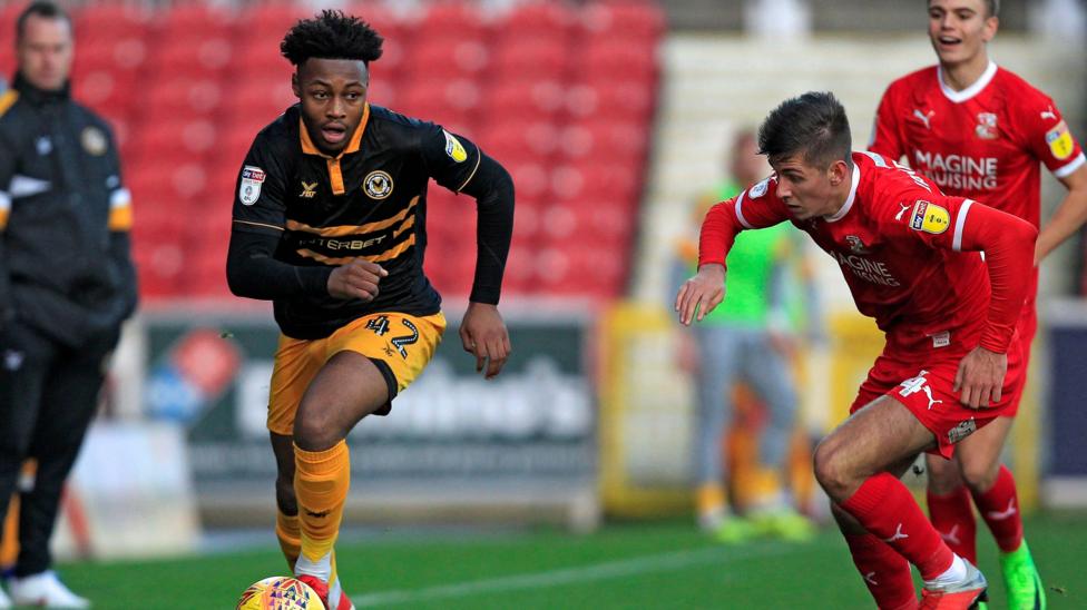 Antoine Semenyo in a black Newport County away kit runs with the ball. Swindon Town players in red are trying to challenge him. Manager Mike Flynn looks on out of focus in the background to the left.