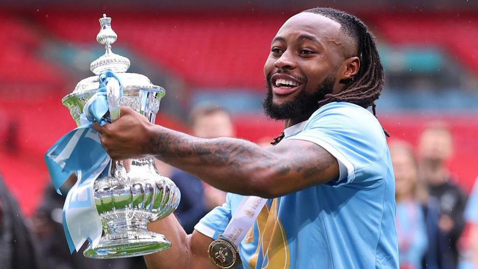 Antoine Semenyo in a sky blue Man City shirt holding the FA Cup trophy at Wembley