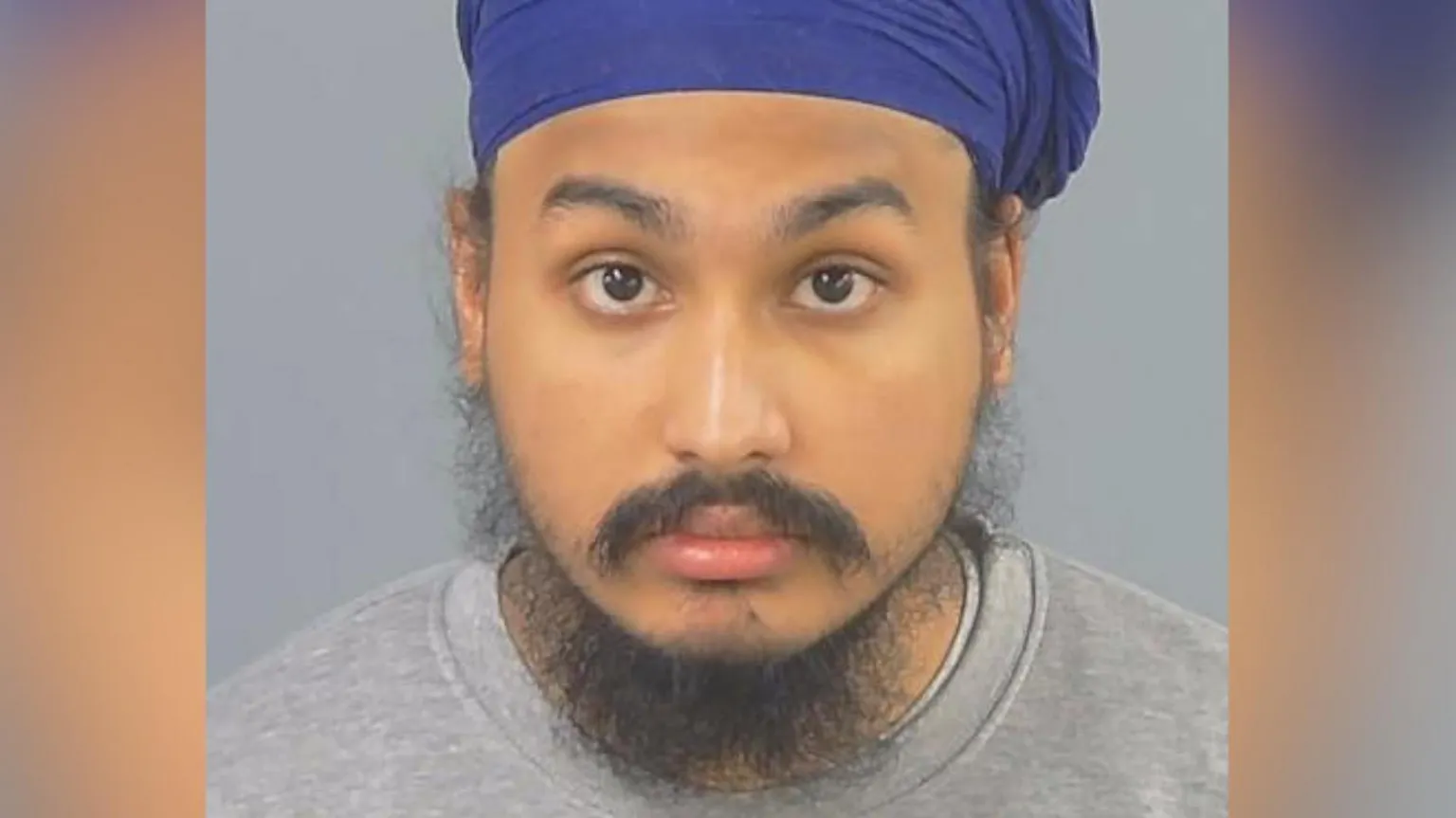 Police custody photograph A portrait photograph of a Sikh man wearing a royal blue turban. He has dark eyes and a moustache and beard. He is wearing a grey sweatshirt. It is a police custody photo.