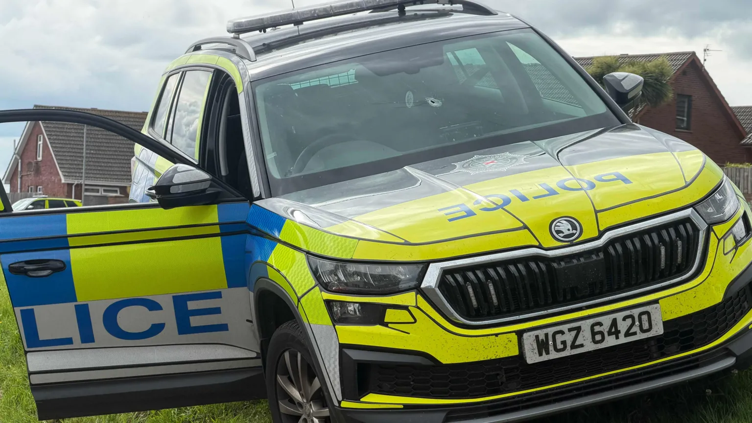 Pacemaker A yellow, blue and white liveried police car with a bullet hole in the windscreen sits on a grassy bank in a housing estate. The driver's door is lying open.
