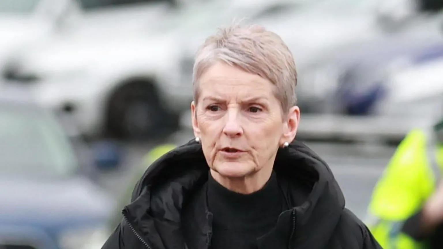  A woman with short cropped grey hair and is wearing a black polo neck top, and a black coat with a hood. She is standing outside, the background is out of focus but the outline of several vehicles can be seen as well as the partial body of someone in a hi-viz jacket
