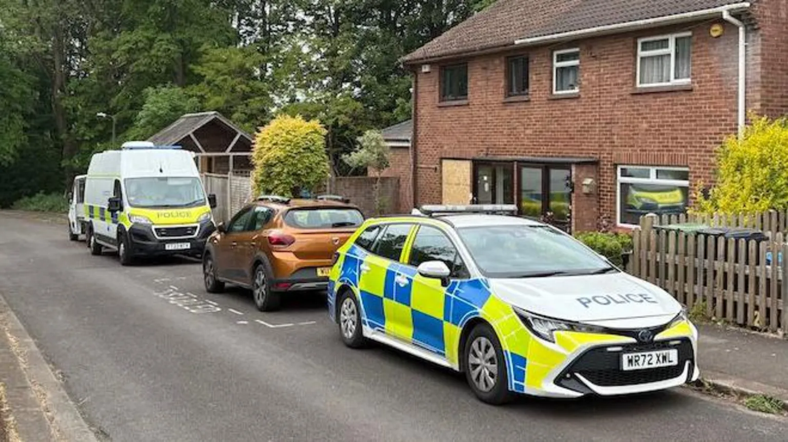 Police cars parked on a residential street. They are outside a house with a boarded up window or door.