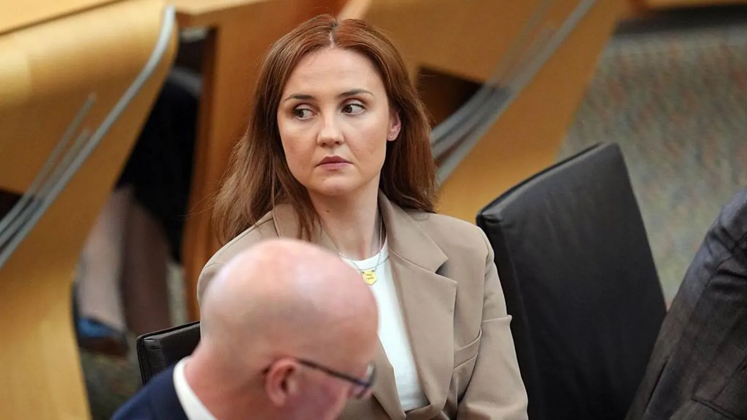 Màiri McAllan wears a beige blazer and white top. She has long brown hair and looks off to the side while sitting next to John Swinney in the Scottish parliament