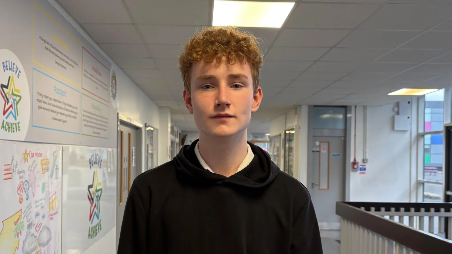 Secondary school pupil Zak. He is wearing a black jumper with a white shirt and has brown curly hair. He is standing in the school corridor, where there are infographics on the wall and doors to classrooms in the background.
