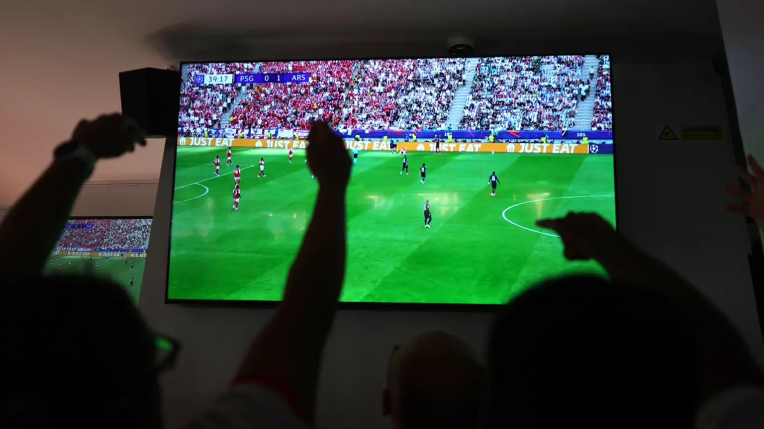  A football match shown on a large TV. Viewers are seen from behind cheering, with their arms in the air, their figures are silhouettes. 