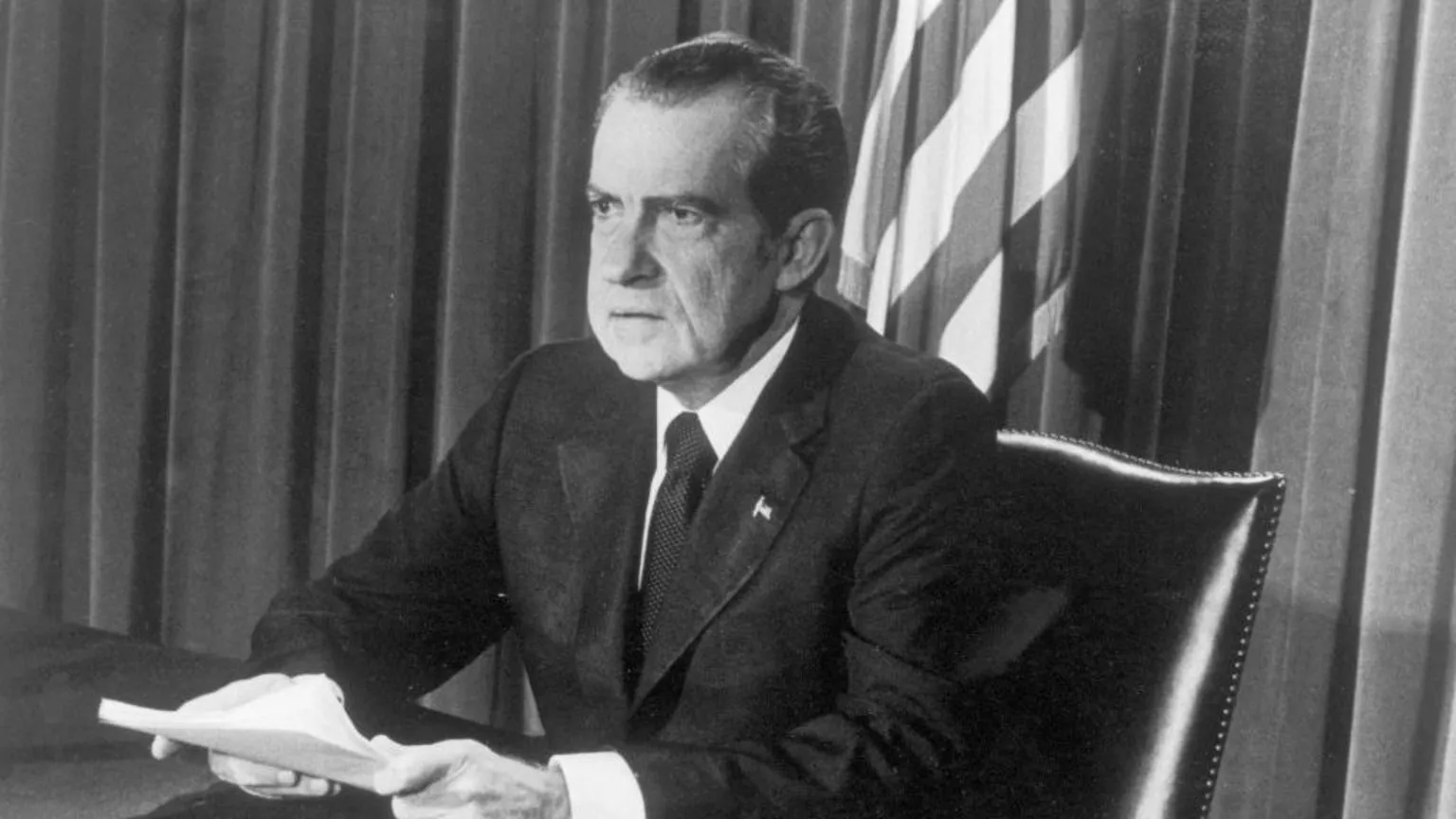 Black and white image of Former US President Richard Nixon taken during live broadcast of resignation in 1974. He is wearing a suit, clutching papers and sitting at a desk.