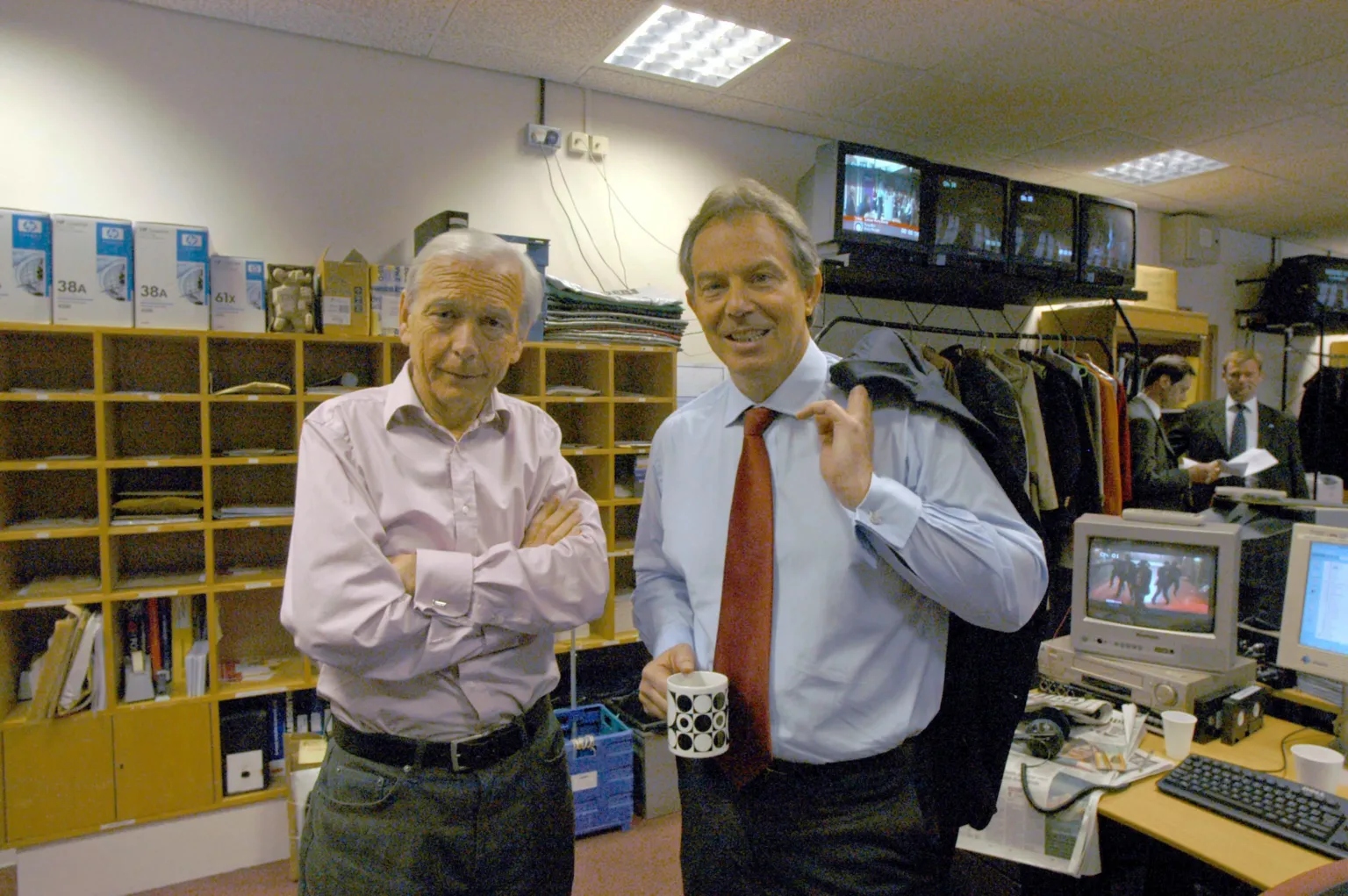 Humphrys - here with former Prime Minster Tony Blair. He crosses his arms, wearing a pink shirt and jeans with a belt. Blair wears a light blue shirt with bronze tie, and holds his blazer over his shoulder. They are standing in an office, with computers in the background.