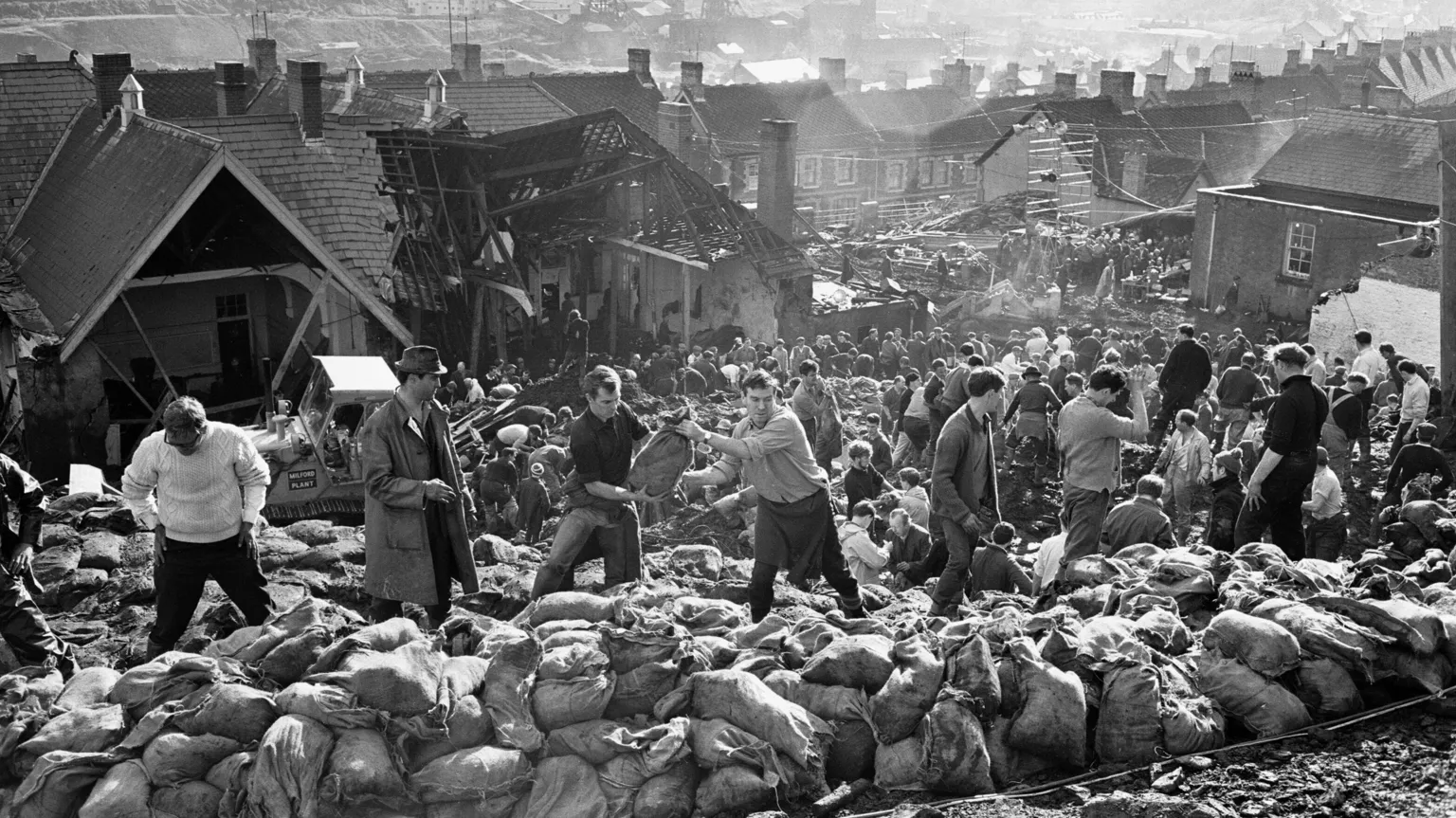 Mirrorpix via A black and white image shows rows of rescue workers moving the coal after the Aberfan disaster, with rows of sandbags in front of them. A damaged building can be seen in the background, with the town of Aberfan behind.